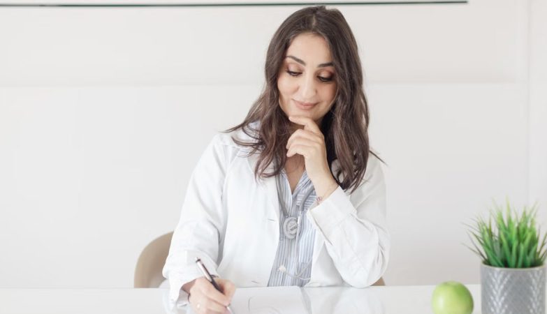 A female nutritionist in a lab coat writing notes with an apple and plant on the desk.
